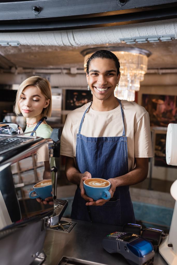Positive african american barista holding cup of coffee near colleague and payment terminals in cafe  - Photo, Image