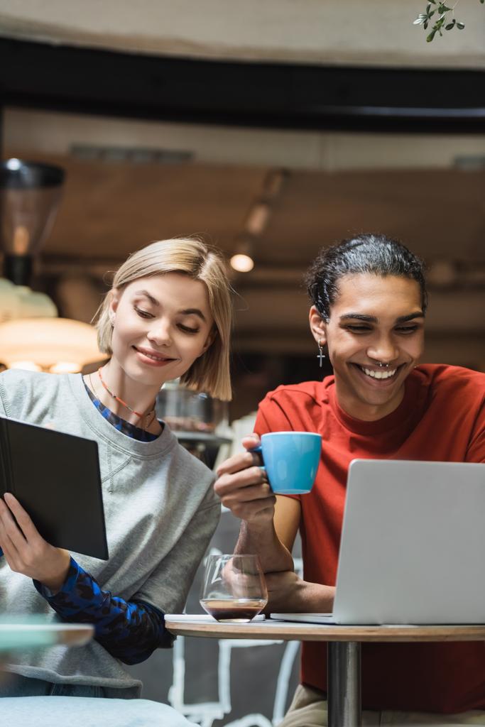 Hymyilevät monikansalliset freelancerit käyttävät kannettavaa tietokonetta ja muistikirjaa kahvilan lähellä  - Valokuva, kuva
