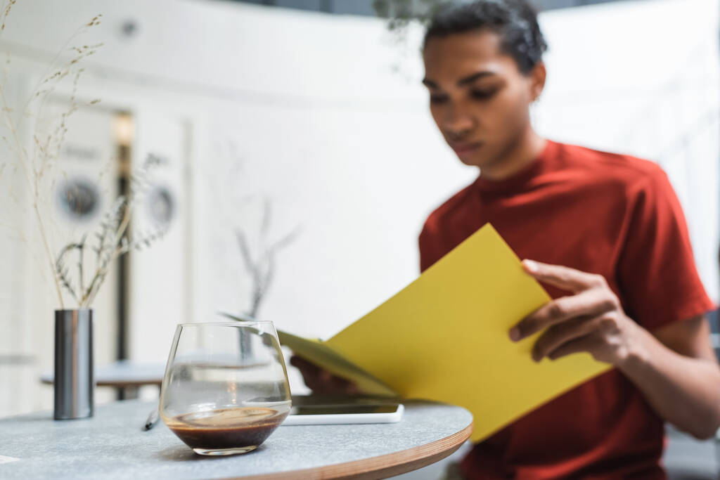 Káva ve skle a smartphone v blízkosti rozmazané africký Američan drží papírové složky v kavárně  - Fotografie, Obrázek