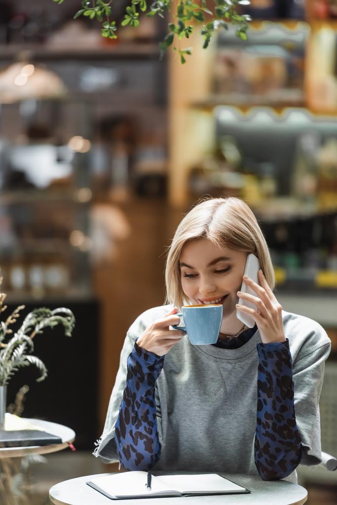 Positieve vrouw praten op smartphone en het drinken van koffie in de buurt van notebook in cafe  - Foto, afbeelding