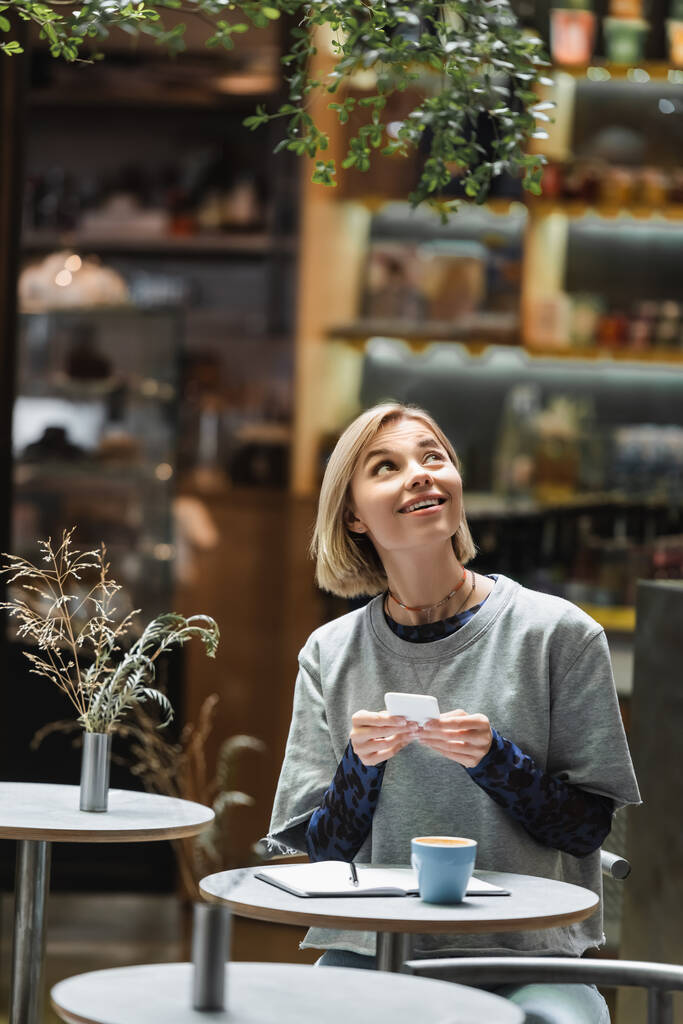 Lächelnde blonde Frau mit Smartphone und Blick nach oben in der Nähe von Kaffee und Notizbuch im Café  - Foto, Bild