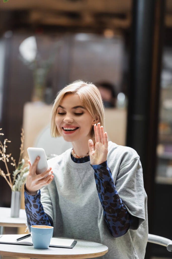 Позитивна жінка має відеодзвінок на смартфон біля кави та ноутбука в кафе
  - Фото, зображення