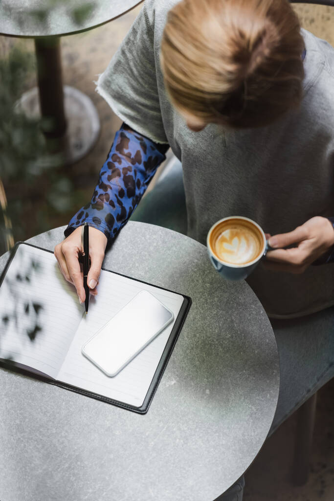 Yleisnäkymä blondi nainen kirjoittaa muistikirjaan lähellä matkapuhelin ja tilalla cup kahvilassa  - Valokuva, kuva