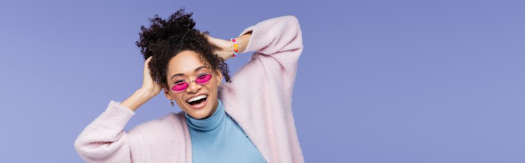heureuse femme afro-américaine dans des lunettes de soleil élégantes ajustant les cheveux bouclés isolés sur violet, bannière - Photo, image
