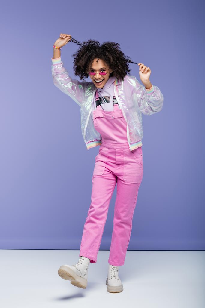 full length of positive african american woman in sunglasses and pink overalls pulling curly hair on purple - Photo, Image