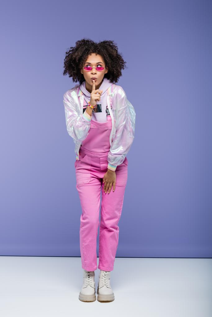 pleine longueur de femme afro-américaine en lunettes de soleil et salopettes montrant signe de silence sur violet - Photo, image