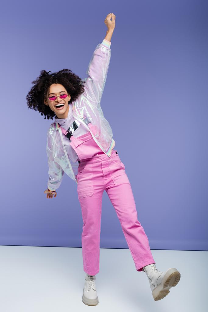 teljes hossza izgatott afro-amerikai nő stílusos overallban és napszemüveg pózol lila - Fotó, kép