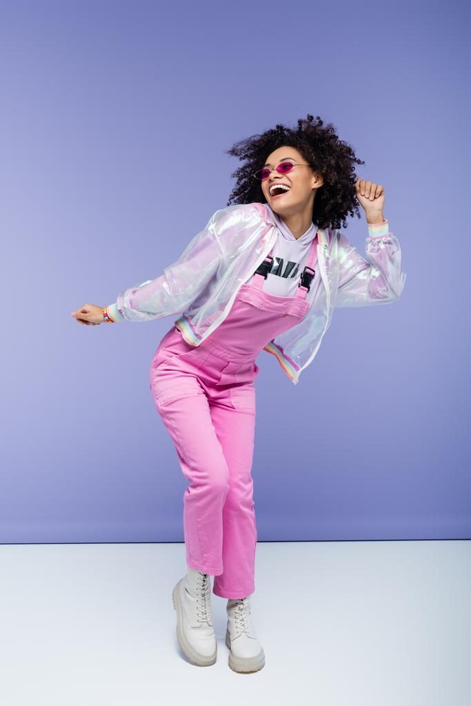 volle Länge der positiven afrikanisch amerikanische Frau in trendigen rosa Overalls posiert auf lila - Foto, Bild