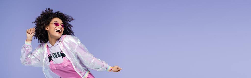 весёлая африканская американка в розовых солнцезащитных очках жестикулирует изолированным на фиолетовом, баннере - Фото, изображение