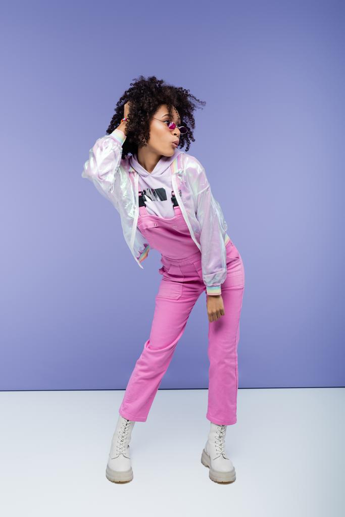 full length of surprised african american woman in stylish overalls posing on purple - Photo, Image