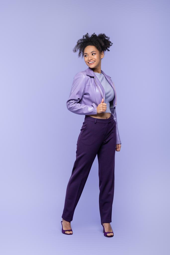 täyspitkä positiivinen ja nuori afrikkalainen amerikkalainen nainen violetti nahkatakki ja housut poseeraa violetti - Valokuva, kuva