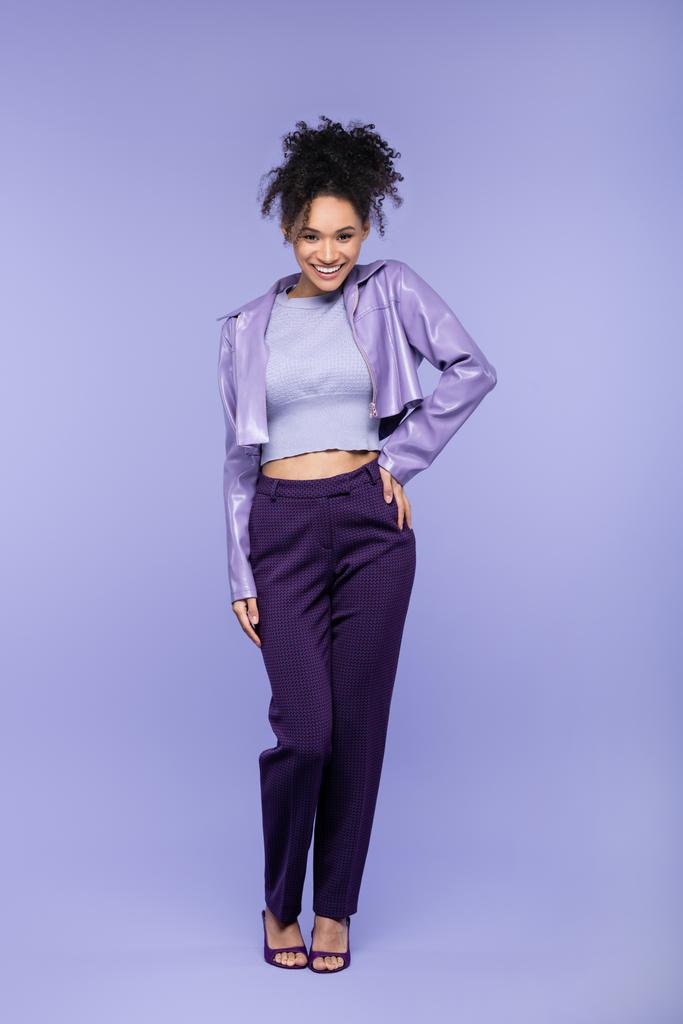 piena lunghezza di allegra donna afroamericana in giacca di pelle viola e pantaloni in posa con mano sul fianco su viola - Foto, immagini