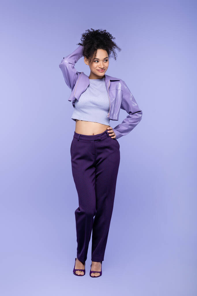 повна довжина кокетливої афроамериканської жінки в фіолетовій шкіряній куртці і штанях позує з рукою на стегні на фіолетовому
 - Фото, зображення