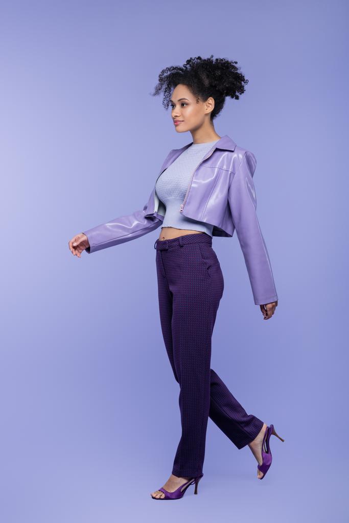 longueur totale de femme afro-américaine bouclée en veste et pantalon en cuir violet marchant sur violet - Photo, image