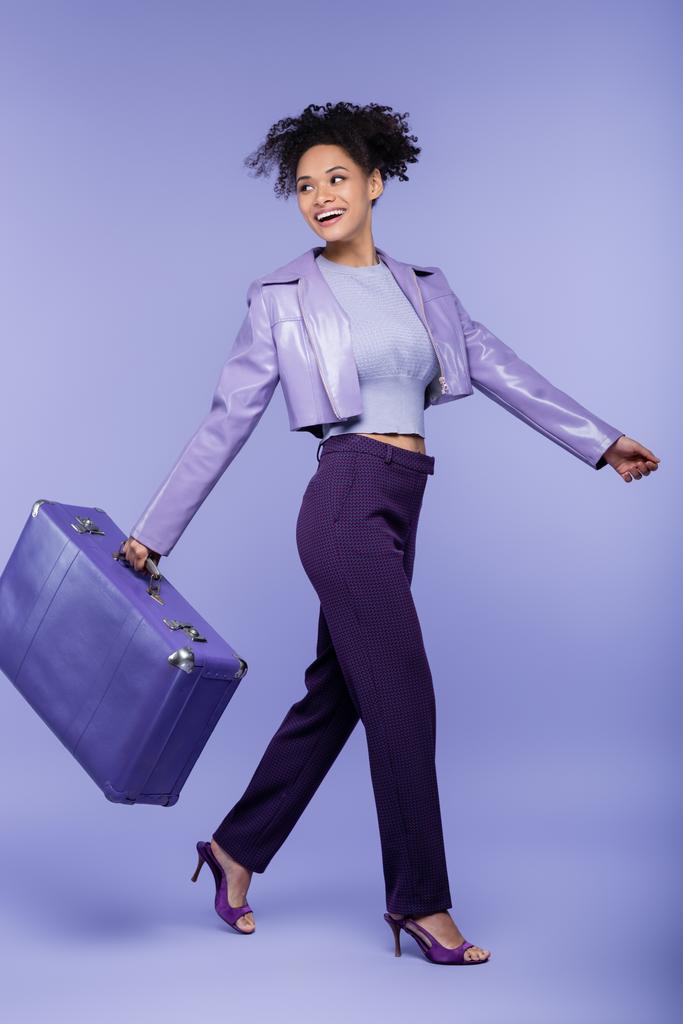 повна довжина щасливої афроамериканської жінки, що йде з валізою на фіолетовому
  - Фото, зображення