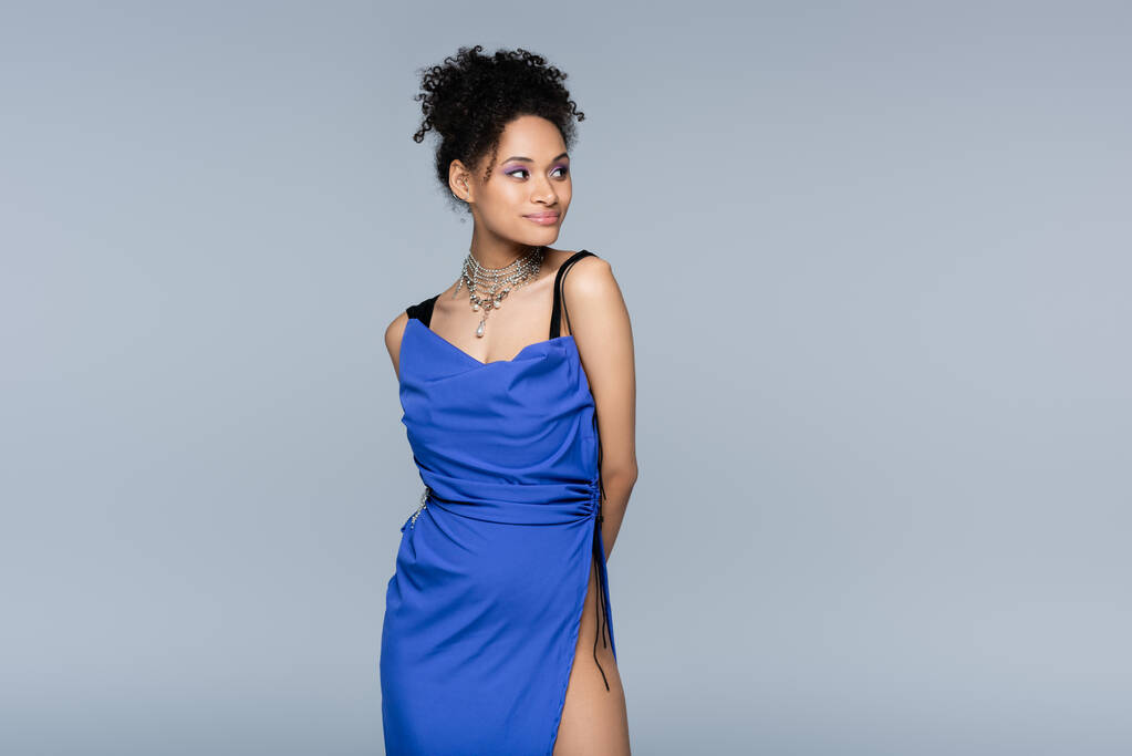 усміхнена афроамериканська жінка в яскраво-блакитній сукні дивиться геть, позує ізольовано на сірому
 - Фото, зображення