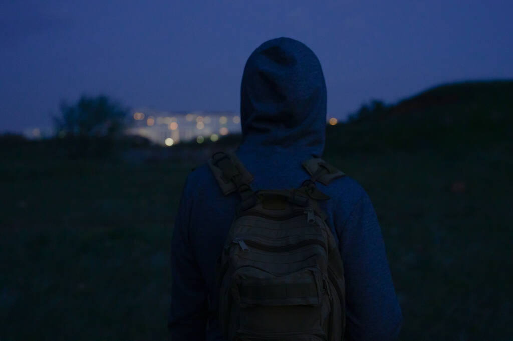 senzatetto in felpa con cappuccio con zaino a guardare sugli edifici notturni della città durante le escursioni al tramonto da solo - Foto, immagini