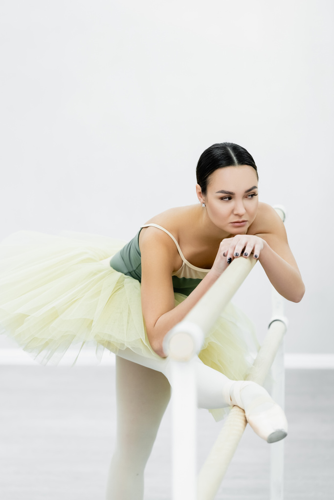 ballerina in tutu stretching at barre while training in studio - Photo, Image