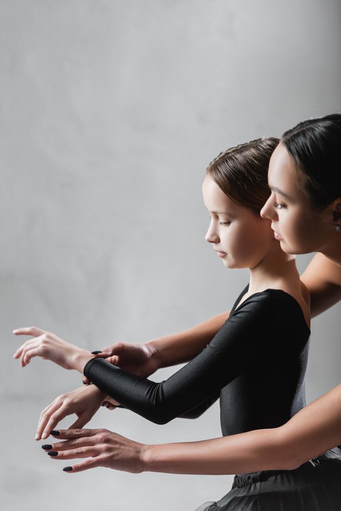 πλαϊνή άποψη του δασκάλου χορού που δείχνει στοιχεία μπαλέτου στο κορίτσι σε γκρι φόντο - Φωτογραφία, εικόνα