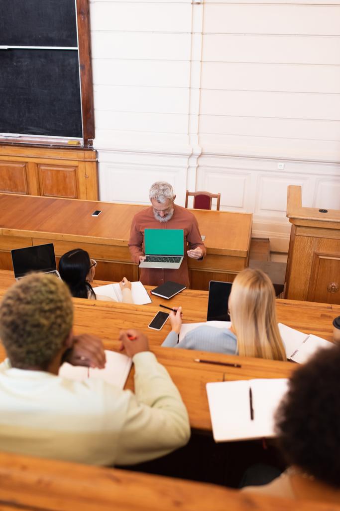 Professeur mature tenant un ordinateur portable avec écran vert près des étudiants multiethniques à l'université  - Photo, image