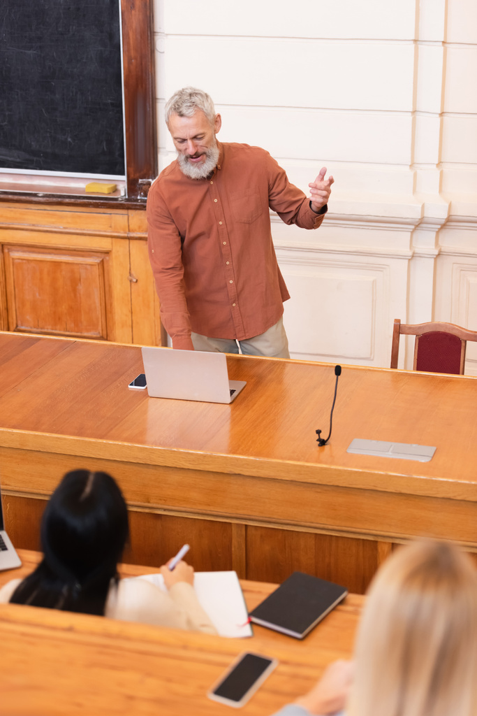 Teacher using laptop near blurred students in university  - Photo, Image