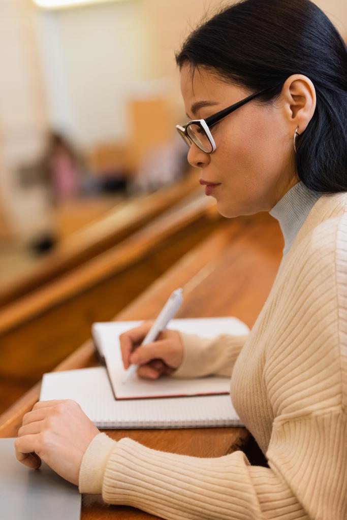 Боковой вид студента, пишущего на размытой тетрадке возле ноутбука в университете  - Фото, изображение