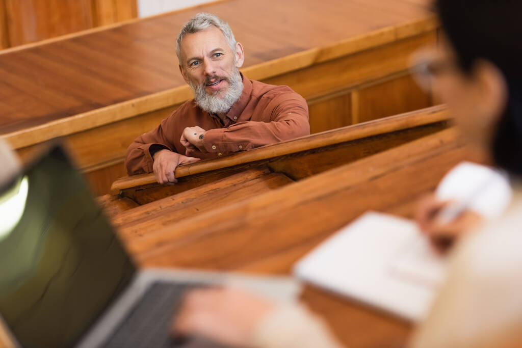 Nauczyciel w średnim wieku patrząc na zamazanego studenta z laptopem podczas wykładu na uniwersytecie  - Zdjęcie, obraz