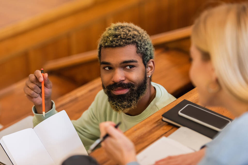Étudiant afro-américain souriant pointant du doigt un ordinateur portable près d'un ami flou à l'université - Photo, image