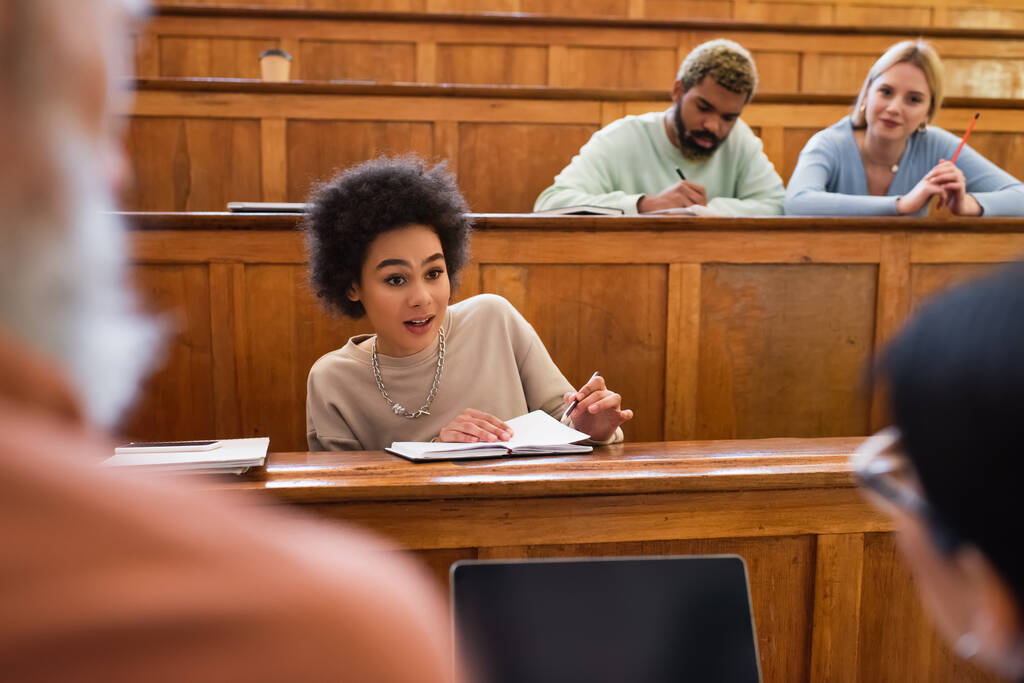Junge afrikanisch-amerikanische Studentin mit Notizbuch im Gespräch mit Lehrer und Freund in der Nähe von Laptop in der Universität  - Foto, Bild