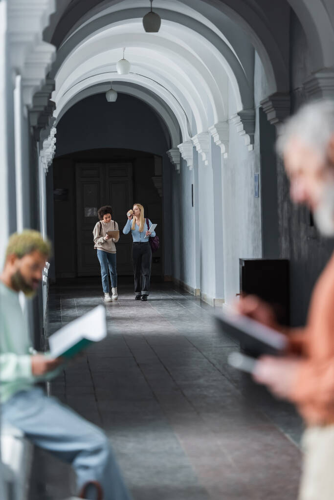 Міжрасові студенти з кавою і ноутбуком ходять в університетському коридорі
  - Фото, зображення