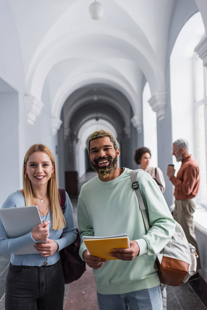 Uśmiechnięci wielonarodowi studenci z notatnikami i laptopem patrzący w kamerę w korytarzu uniwersyteckim  - Zdjęcie, obraz