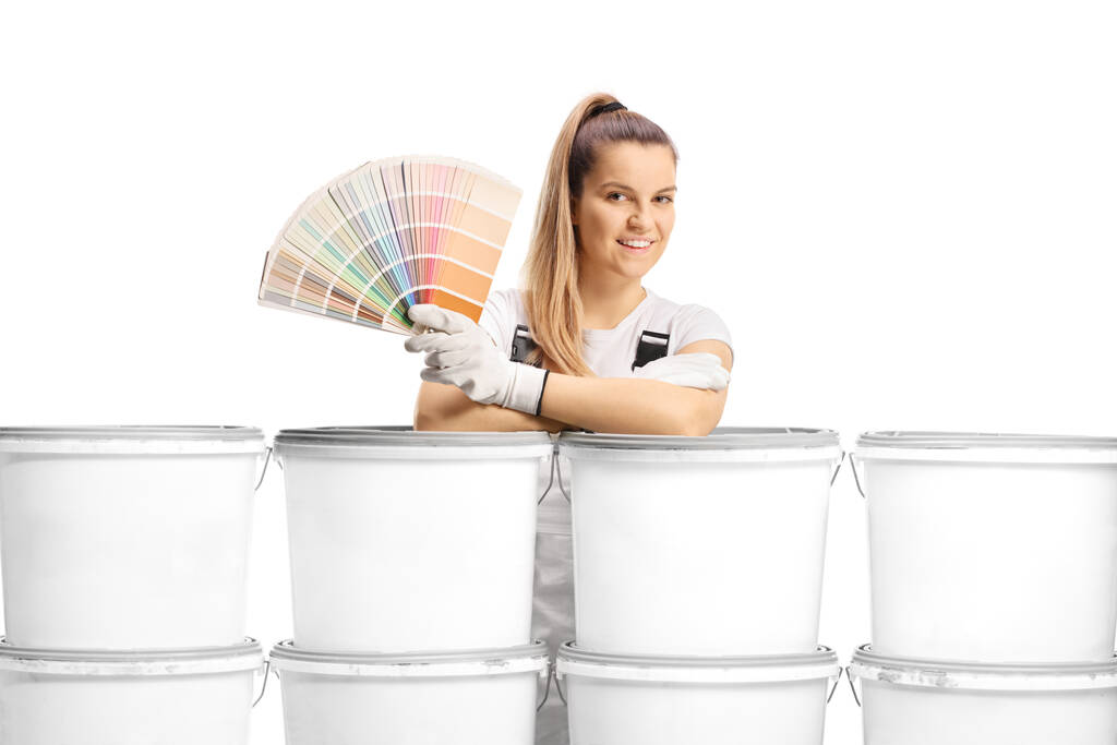 Γυναίκα ζωγράφος σπίτι ακουμπά σε ένα σωρό κουβάδες και κρατώντας μια παλέτα χρωμάτων που απομονώνονται σε λευκό φόντο - Φωτογραφία, εικόνα