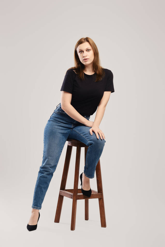 Young woman in black t shirt and jeans posing on tall wooden chair over grey background - Photo, Image