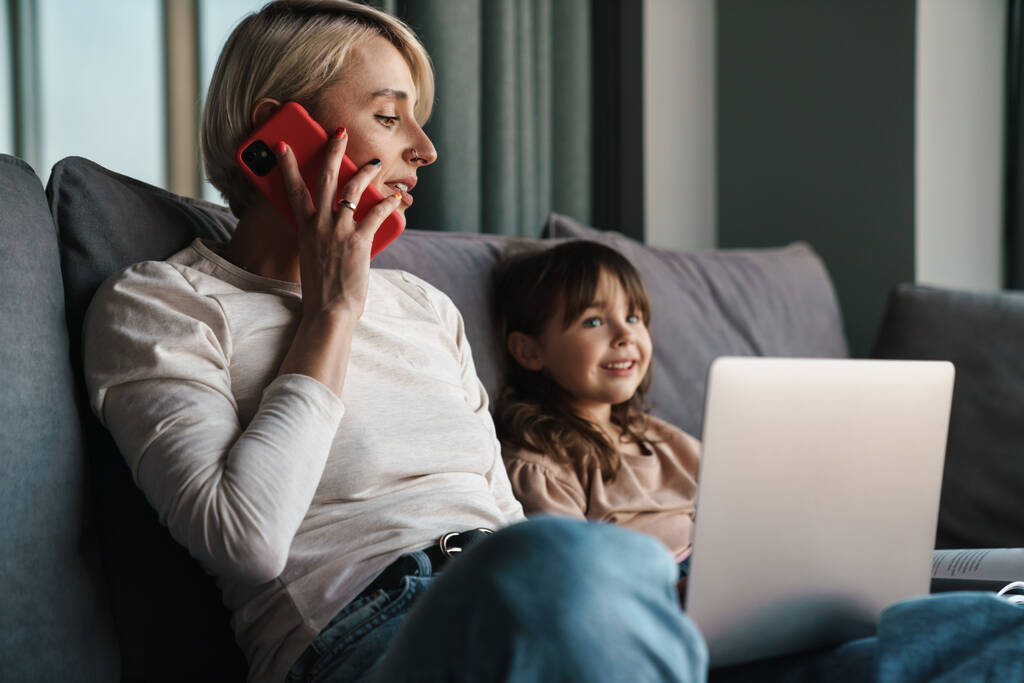 Šťastná mladá matka a její dcera sedí na gauči s notebookem, dívá se na obrazovku, na videohovor nebo sleduje filmy, žena mluví na mobilním telefonu - Fotografie, Obrázek