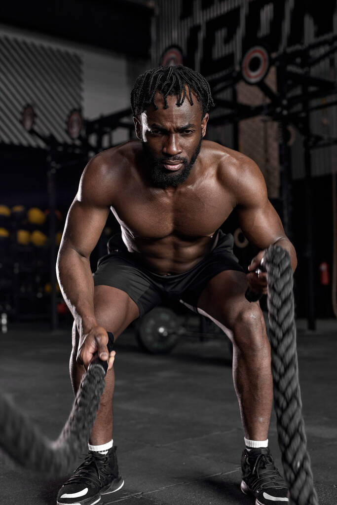 Hombre africano joven haciendo entrenamiento de fuerza usando cuerdas de batalla en el moderno gimnasio oscuro. - Foto, imagen