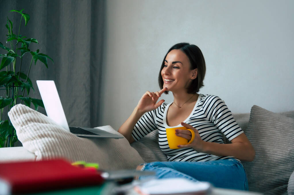 Zamknij zdjęcie pięknej uśmiechniętej freelancerki pracującej na laptopie, leżącej na dużej kanapie i pijącej kawę w domu - Zdjęcie, obraz