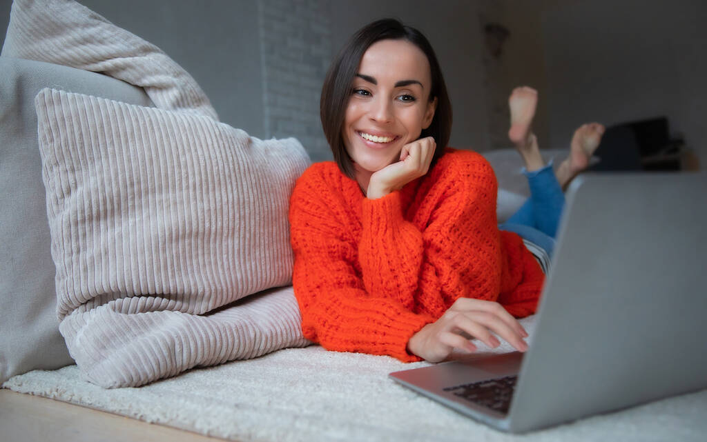 Όμορφη νεαρή ευτυχισμένη κοπέλα που βρίσκεται στο πάτωμα του σπιτιού και εργάζονται για το laptop ή surfing στο δίκτυο - Φωτογραφία, εικόνα