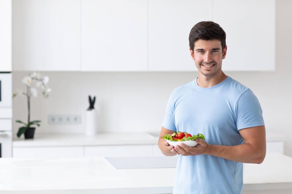 Молодой красивый улыбающийся мужчина на кухне с овощным салатом в руках, хорошо смотрится в голубой футболке. Концепция здорового и вегетарианского питания. - Фото, изображение