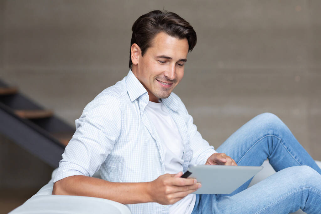 Bonito homem sorridente com tablet digital no sofá em casa, compras on-line e net surf. - Foto, Imagem