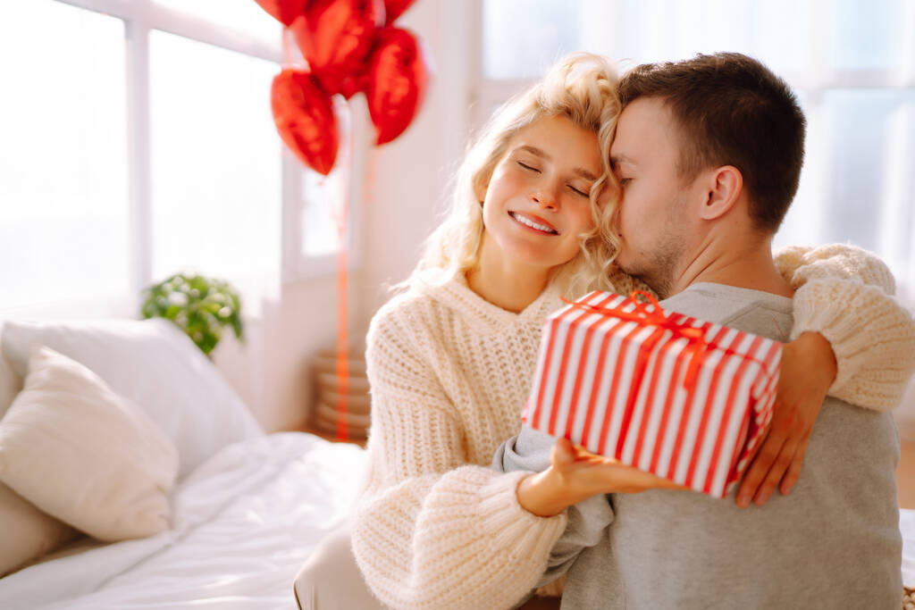 Schönes junges Paar zu Hause feiert den Valentinstag. Liebende beschenken sich gegenseitig. Umarmen, küssen und die gemeinsame Zeit genießen. - Foto, Bild