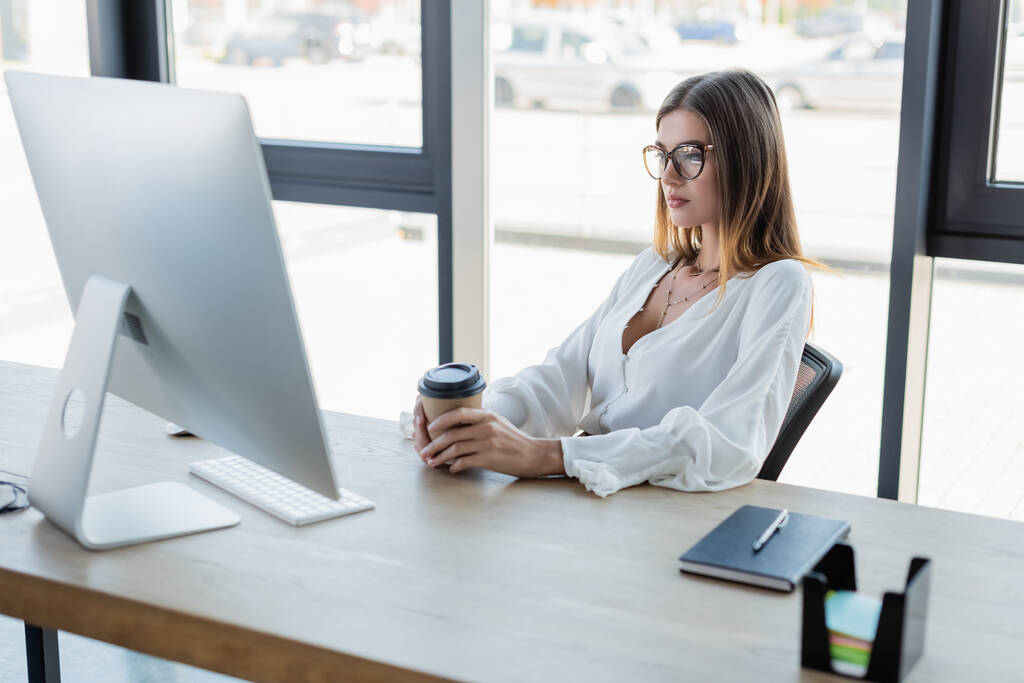 businesswoman in glasses holding paper cup and looking at computer monitor in office - Photo, Image