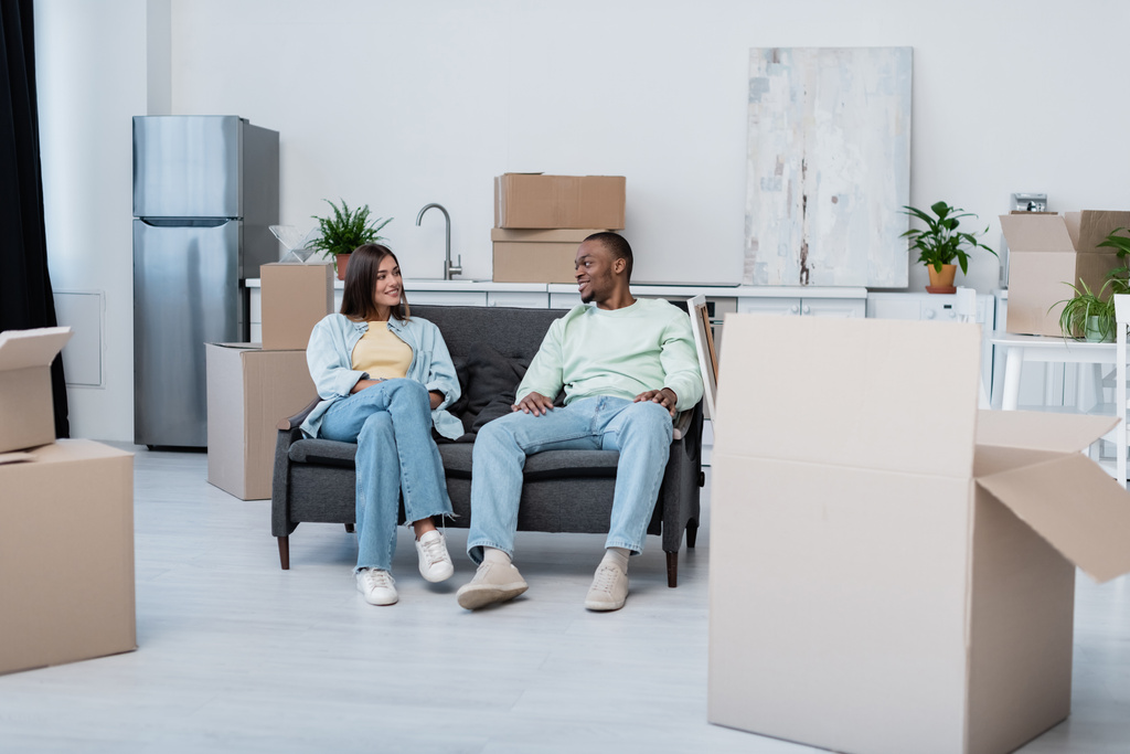 happy interracial couple sitting on sofa around carton boxes during relocation  - Photo, Image