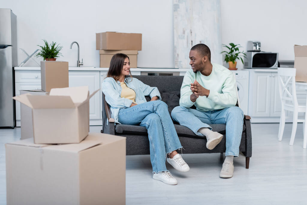 cheerful interracial couple looking at each other while sitting on sofa near carton boxes during relocation  - Photo, Image
