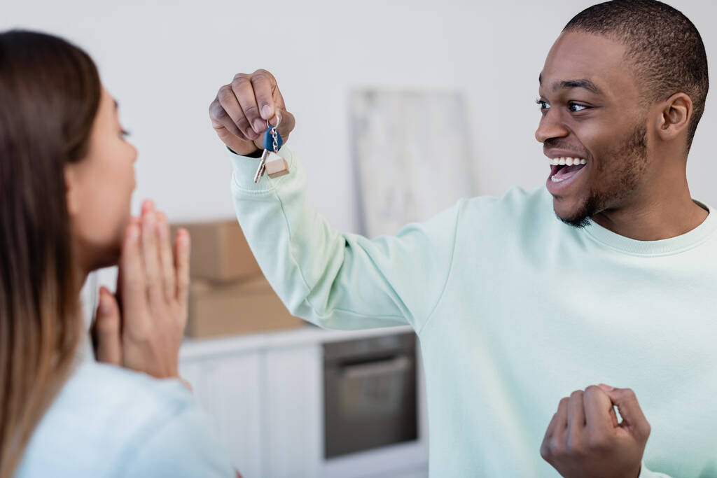 Mutlu Afrikalı Amerikalı adam yeni dairede bulanık kız arkadaşının yanında anahtarı tutuyor. - Fotoğraf, Görsel