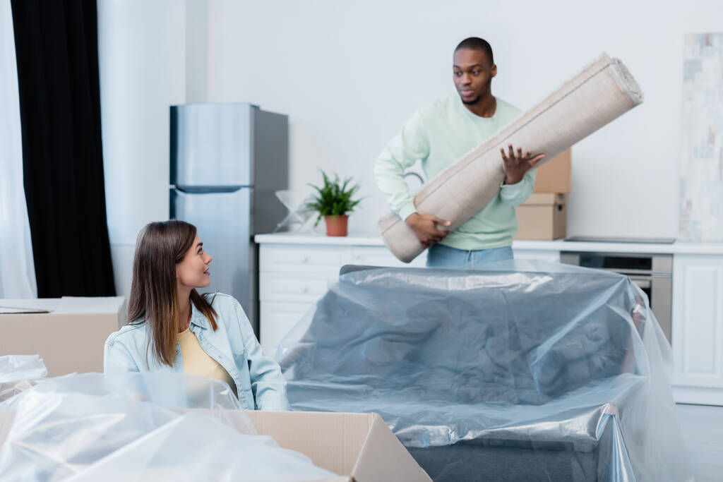 cheerful woman looking at african american boyfriend holding rolled carpet in new home - Photo, Image