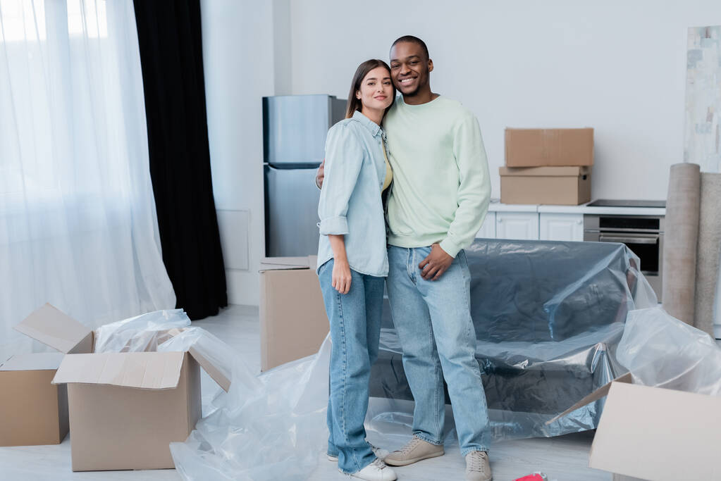 cheerful interracial couple hugging while standing in new apartment  - Photo, Image