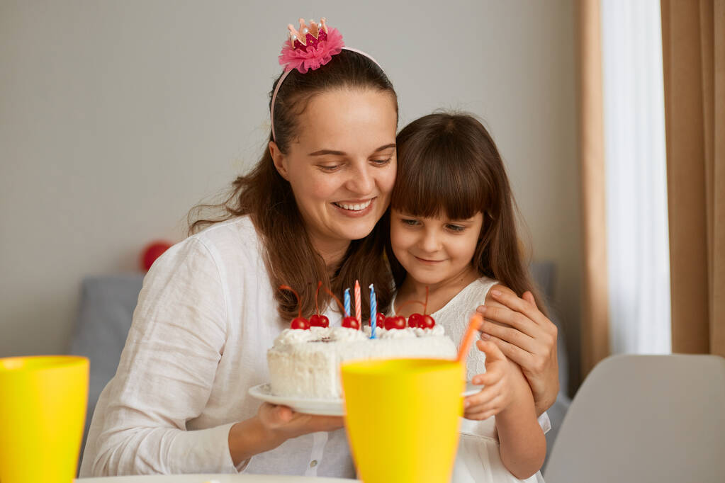 Poziome ujęcie szczęśliwej rodziny, matki i córki siedzących przy stole i przytulających się, świętujących urodziny, wyrażających optymistyczne emocje, mała dziewczynka zdmuchnie świeczkę. - Zdjęcie, obraz