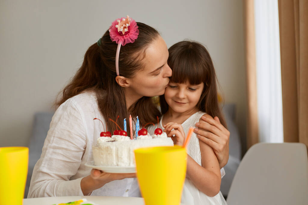 Εσωτερική φωτογραφία γυναίκας με θηλυκό παιδί να γιορτάζει τα γενέθλιά της, μητέρα να φιλάει την κορούλα της, να συγχαίρει το παιδί με τα γενέθλιά της, να έχει μέρος στο σπίτι. - Φωτογραφία, εικόνα