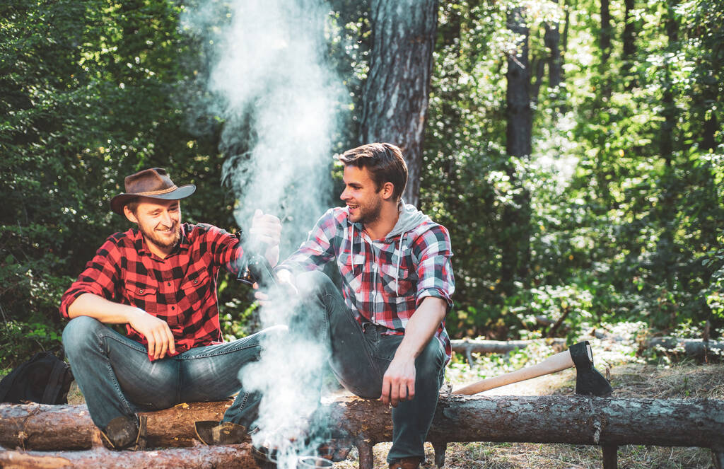 Friends relaxing near campfire after day hiking or gathering mushrooms. Company two male friends enjoy relaxing together in forest. Two friends guy camping in forest. - Photo, Image