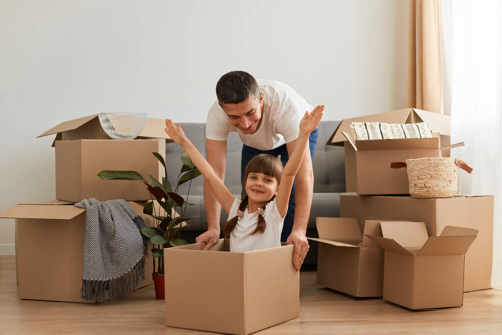 Hravý otec se baví ve svém novém bytě v den stěhování, šťastný muž a jeho dcera hrají spolu s kartonovou krabicí při stěhování, dívka roztahuje ruce, slaví stěhování. - Fotografie, Obrázek